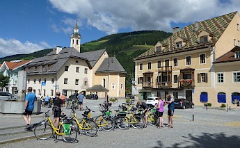 Cyclists gathered with their bikes in a town square in the Dolomites, Italy, with colorful buildings and a church in the background.
