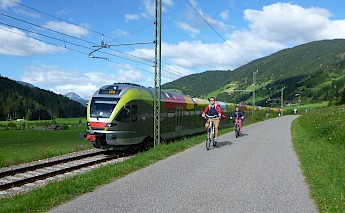 Two cyclists riding along a path next to a colorful train in a green valley in the Dolomites, Italy.