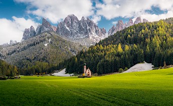 A small church stands in a lush green meadow, with the jagged peaks of the Dolomite Mountains rising in the background under a partly cloudy sky.