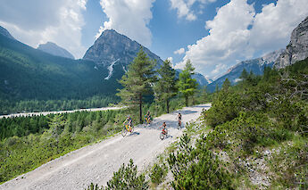 Cyclists riding on a gravel path in the Dolomites with a view of the mountains and lush valley below.