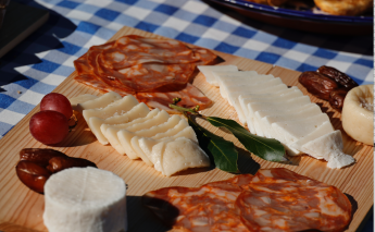 A wooden platter with sliced cheeses, cured meats, grapes, and dates on a blue checkered tablecloth.