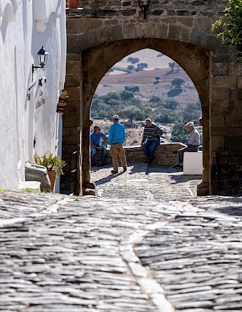 A cobblestone path in Monsaraz, Portugal, framed by an archway, with a few people gathered and hills in the background.