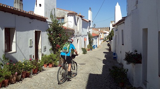 A cyclist on a cobblestone street lined with whitewashed houses and colorful flowers in Portugal.