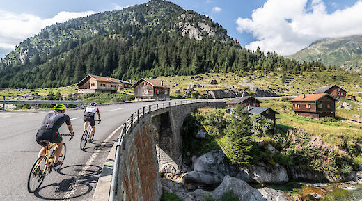 Cyclists riding on a scenic road with traditional wooden houses and a forested mountain in the background.