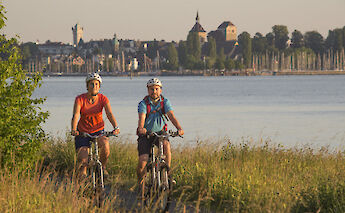 Two cyclists riding along a lakeside path with a scenic view of historic buildings in the background.