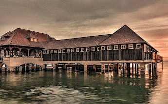 A wooden structure on stilts extends over a peaceful body of water at sunset, with a warm glow in the sky.