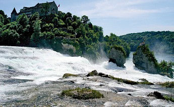Rhine Falls with mist rising from the cascading water, surrounded by green hills and a building on the hilltop.