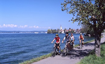 Cyclists riding alongside a large body of water with a church and small town in the background.