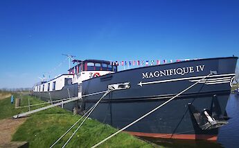 A boat named Magnifique IV is docked with flags adorning the deck against a clear blue sky.