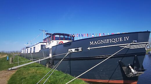 A boat named Magnifique IV is docked with flags adorning the deck against a clear blue sky.