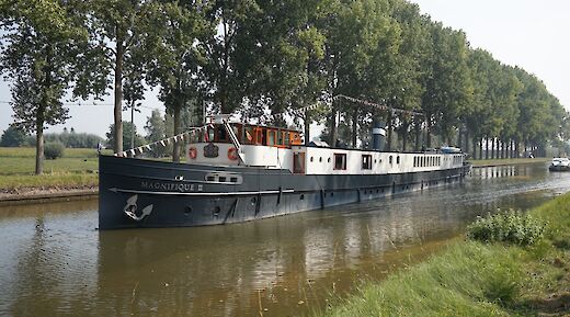 A boat named Magnifique II navigating a canal lined with trees.