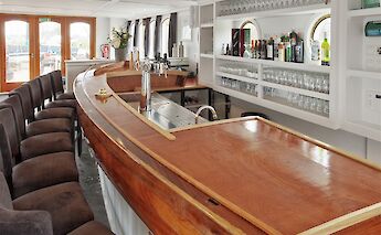 A bar with a wooden counter, stools, and shelves stocked with glasses and bottles.