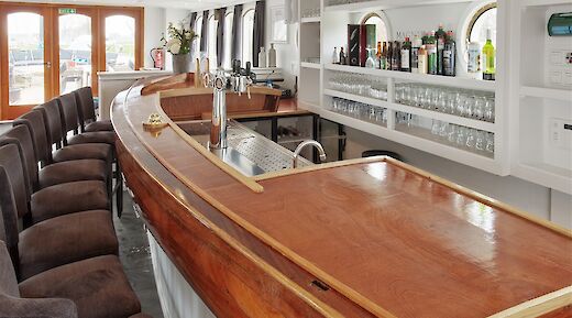 A bar with a wooden counter, stools, and shelves stocked with glasses and bottles.