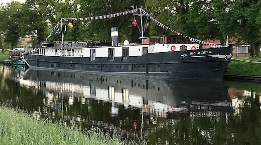 The Magnifique III vessel moored along a canal, with colorful flags strung across its deck amid green trees.