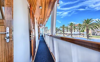 A narrow walkway outside ship cabins with wooden doors and a view of palm trees and buildings along…