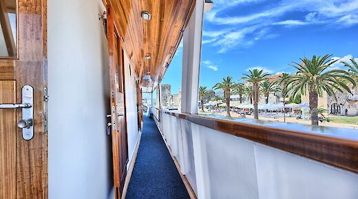 A narrow walkway outside ship cabins with wooden doors and a view of palm trees and buildings along the shore.