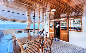 A covered deck on a yacht featuring a wooden table and chairs, with a view of the sea in the…