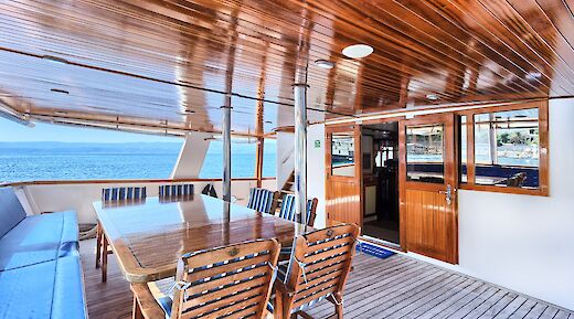 A covered deck on a yacht featuring a wooden table and chairs, with a view of the sea in the background.