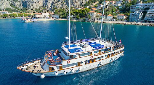 Aerial view of a white yacht with blue awnings named Romantica sailing near a coastal town with mountainous terrain in the background.