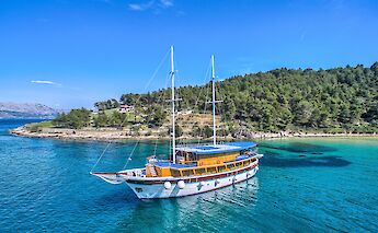 Aerial view of the San Snova sailing near a green, forested island in clear blue waters.