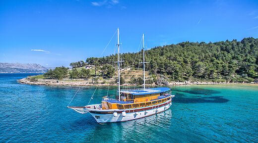 Aerial view of the San Snova sailing near a green, forested island in clear blue waters.