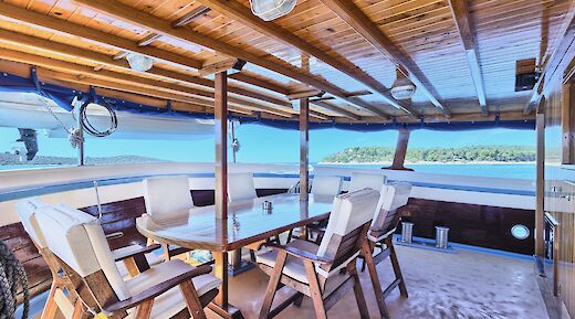 Covered lower deck area with wooden table and cushioned chairs on the San Snova, with views of the sea and a distant green shoreline.