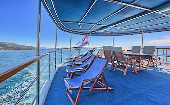 Upper deck of the San Snova with lounge chairs and a table under a canopy, overlooking the sea with…