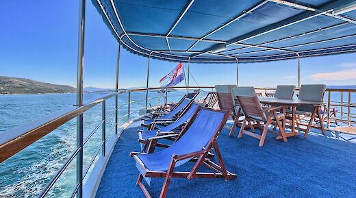 Upper deck of the San Snova with lounge chairs and a table under a canopy, overlooking the sea with a flag in the background.