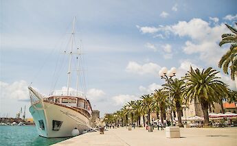 A white ship named Harmonia docked along a promenade lined with palm trees under a clear blue sky.
