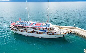 A two-masted boat named Harmonia is docked at a pier, with calm blue waters surrounding it under a…