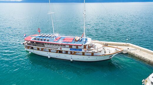 A two-masted boat named Harmonia is docked at a pier, with calm blue waters surrounding it under a clear sky.