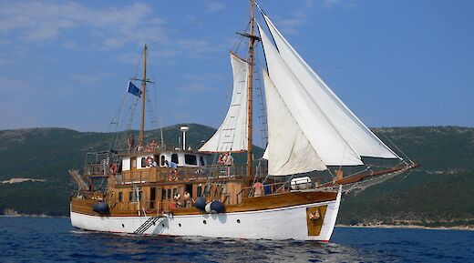 A traditional wooden sailboat named Panagiota with sails unfurled, sailing on the sea against a backdrop of hilly terrain.