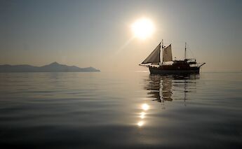 A sailboat named Panagiota silhouetted against the sun on calm waters, with distant mountains in&hellip;