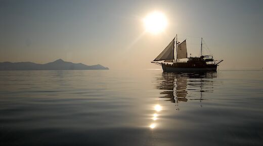 A sailboat named Panagiota silhouetted against the sun on calm waters, with distant mountains in the background.