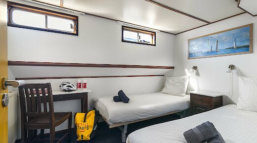 A cozy cabin with two single beds, a small desk with a chair, and a window. Cycling gear, including a helmet and water bottles, is on the desk, and a colorful painting hangs on the wall.