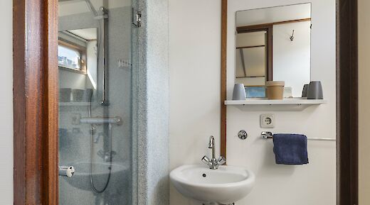 A compact bathroom featuring a sink, mirror, and shower with a glass door. The setting has wooden trim and a small shelf holding toiletries and towels.