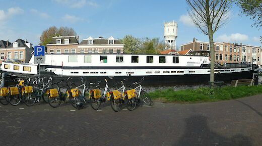 A white boat is moored along a canal with a row of bicycles parked in front. The backdrop includes traditional Dutch-style buildings and a water tower.