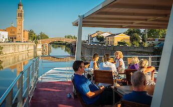 Passengers on the upper deck of Ave Maria enjoying a view of a river with a historic church tower…