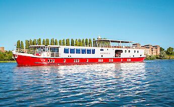 A red and white boat named Ave Maria sailing on a river with trees and historic buildings in the…