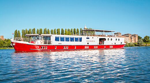 A red and white boat named Ave Maria sailing on a river with trees and historic buildings in the background.