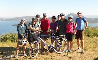 A group of cyclists in helmets poses on a hillside overlooking a scenic lake and distant mountains…