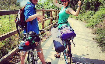 Two cyclists wearing helmets pose on a paved path surrounded by greenery, with their bikes loaded&hellip;