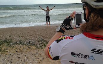 A person wearing cycling gear takes a photograph of another person standing on a sandy beach with…