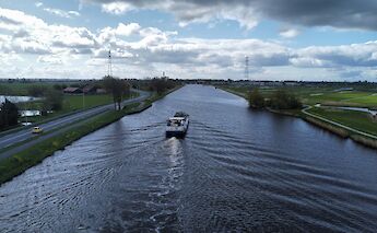 A boat travels along a wide canal under a partly cloudy sky, with fields and roads on either side.