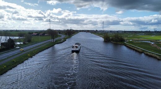 A boat travels along a wide canal under a partly cloudy sky, with fields and roads on either side.