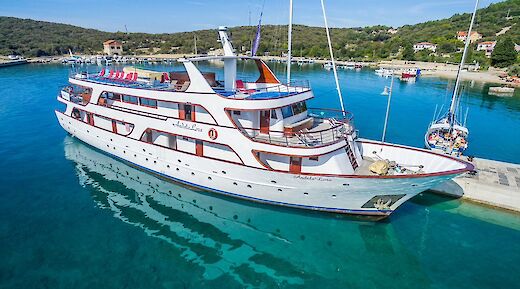 The Andela Lora is docked in clear blue water with surrounding greenery and a few buildings nearby.