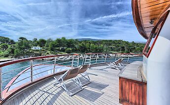 A curved wooden deck area on the Andela Lora boat with sun loungers overlooking a coastline with&hellip;