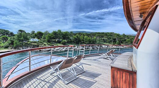 A curved wooden deck area on the Andela Lora boat with sun loungers overlooking a coastline with trees and clear skies.