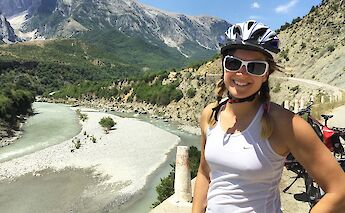 A person wearing a helmet and sunglasses stands smiling by a river with mountainous terrain in the…