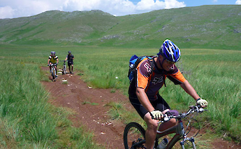 Cyclists riding mountain bikes through a grassy, hilly landscape under a blue sky.